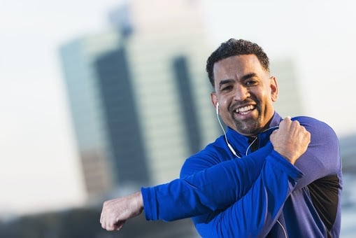 A smiling African American man exercising outdoors, stretching his arms. He is a mature man, in his 40s, facial hair. He is wearing a royal blue and black sweatshirt, with an earbud in one ear, listening to music as he stays in shape. He is in a city, with buildings out of focus behind him.